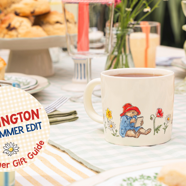 A cream mug of tea with a floral summery design on a picnic tablescape, scones and marmalade in the background. Paddington Summer Edit Gift Guide cream gingham badge sits in the bottom left