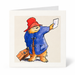 A square greeting card featuring a watercolor illustration of Paddington Bear wearing his signature red hat and blue duffle coat, holding an orange suitcase and a white sheet of paper, on a white background.