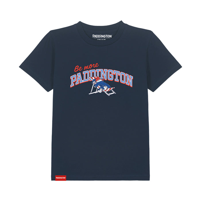 A navy blue short-sleeve t-shirt with a 'Be more PADDINGTON' graphic on the chest, featuring a bear on a deckchair illustration and a red Paddington brand tag near the bottom hem, shown on a plain white background.