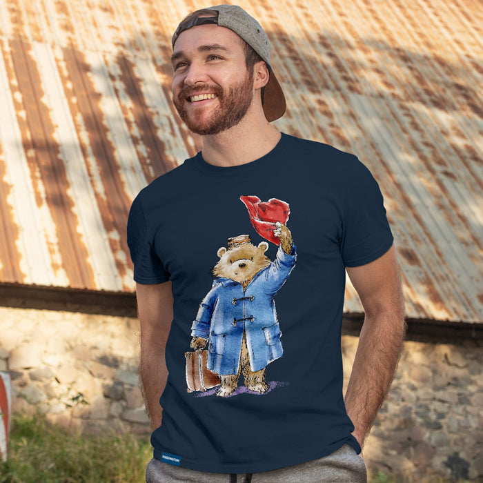 A man wearing a navy blue t-shirt with a colorful graphic of Paddington Bear holding a red hat and suitcase, posed in front of a rustic corrugated metal roof background.
