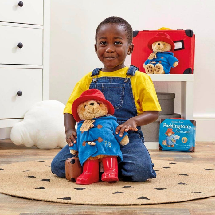 A young child kneeling on a patterned rug in a playroom, holding a Paddington Bear plush toy. A red suitcase, a cloud-shaped cushion, and a Paddington book are visible in the background.