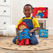 A young child kneeling on a patterned rug in a playroom, holding a Paddington Bear plush toy. A red suitcase, a cloud-shaped cushion, and a Paddington book are visible in the background.