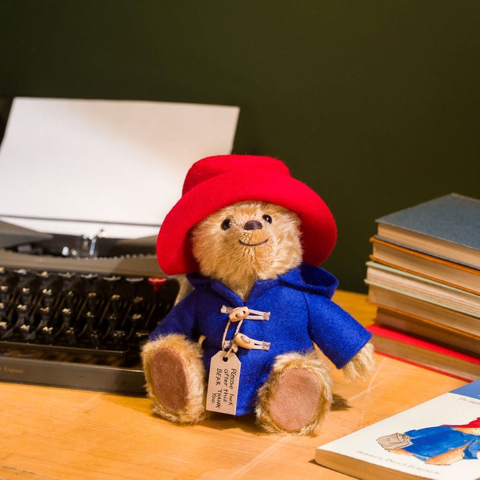 Paddington Teddy bear in a red hat and blue coat on a wooden surface with books and a typewriter in the background.