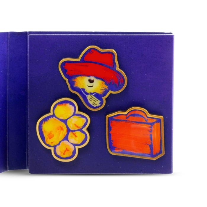 Set of colorful pins featuring a bear with a red hat, paw print, and orange suitcase on a blue card.