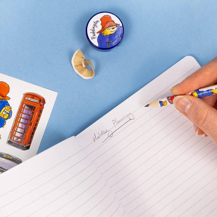 A hand holds a colorful pencil with Padding Bear illustrations while writing on a lined notepad. A matching circular pencil sharpener with Padding Bear is visible on a blue background, along with pencil shavings and a printed sticker sheet.