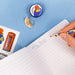 A hand holds a colorful pencil with Padding Bear illustrations while writing on a lined notepad. A matching circular pencil sharpener with Padding Bear is visible on a blue background, along with pencil shavings and a printed sticker sheet.