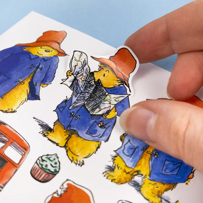 A close-up of a hand peeling a die-cut sticker from a sheet of colorful illustrated stickers. The sticker shows Paddington Bear in a blue duffel coat and orange hat reading a newspaper, with other Paddington-themed stickers visible on the sheet.