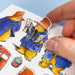 A close-up of a hand peeling a die-cut sticker from a sheet of colorful illustrated stickers. The sticker shows Paddington Bear in a blue duffel coat and orange hat reading a newspaper, with other Paddington-themed stickers visible on the sheet.