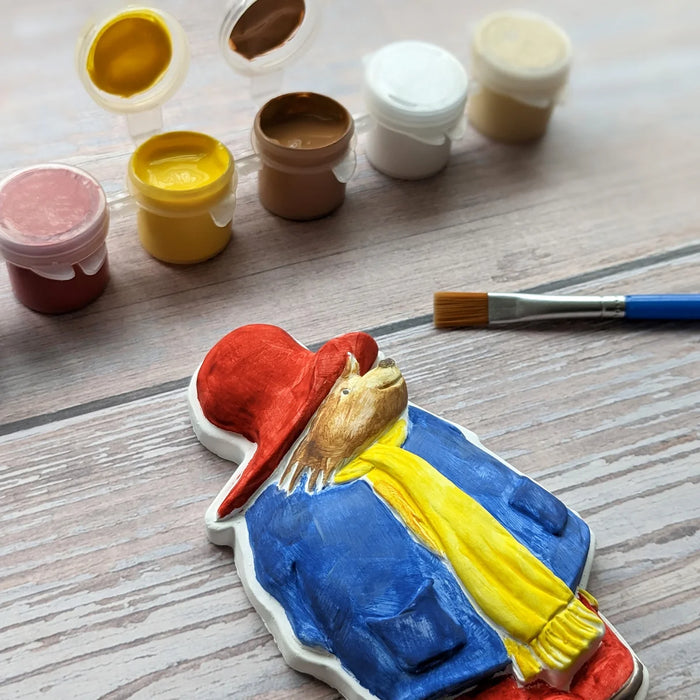 A hand-painted plaster figurine of Paddington Bear, styled on a light wood surface with a paintbrush and small paint pots in the background.