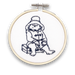 A mini embroidery kit depicting the character Paddington Bear sitting and appear to be enjoying a sandwich.