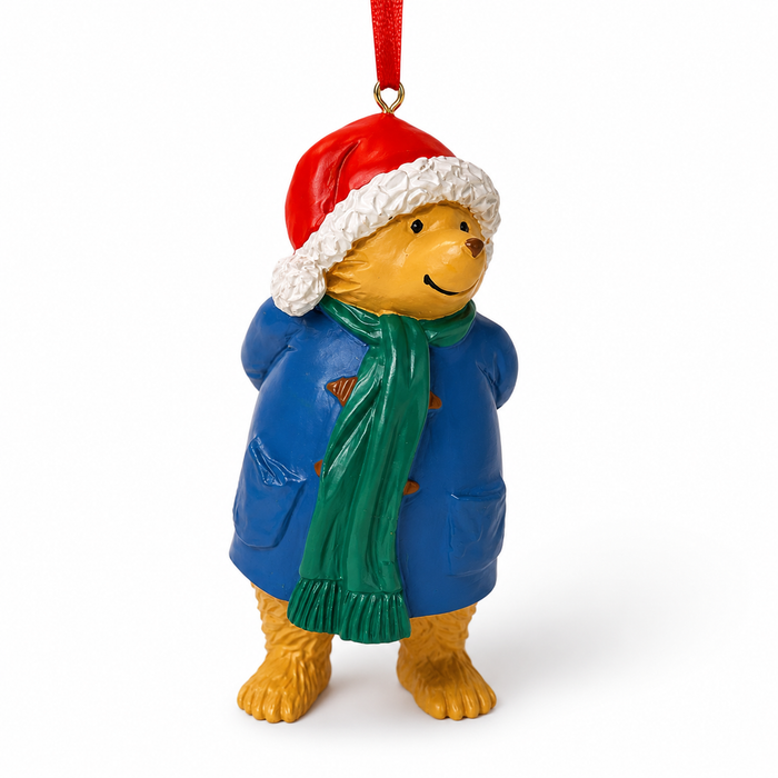 A Pattington bear Christmas ornament wearing a red Santa hat with white trim, a blue coat, and a green scarf, hanging by a red ribbon on a white background.