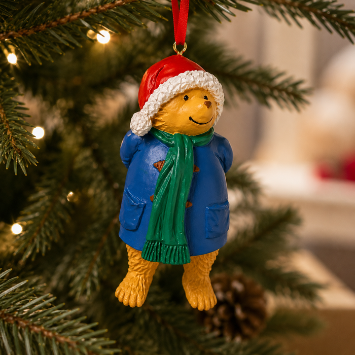 A Paddington bear Christmas ornament hanging from a red ribbon on a decorated Christmas tree. The bear is wearing a red Santa hat with white trim, a blue coat, and a green scarf.