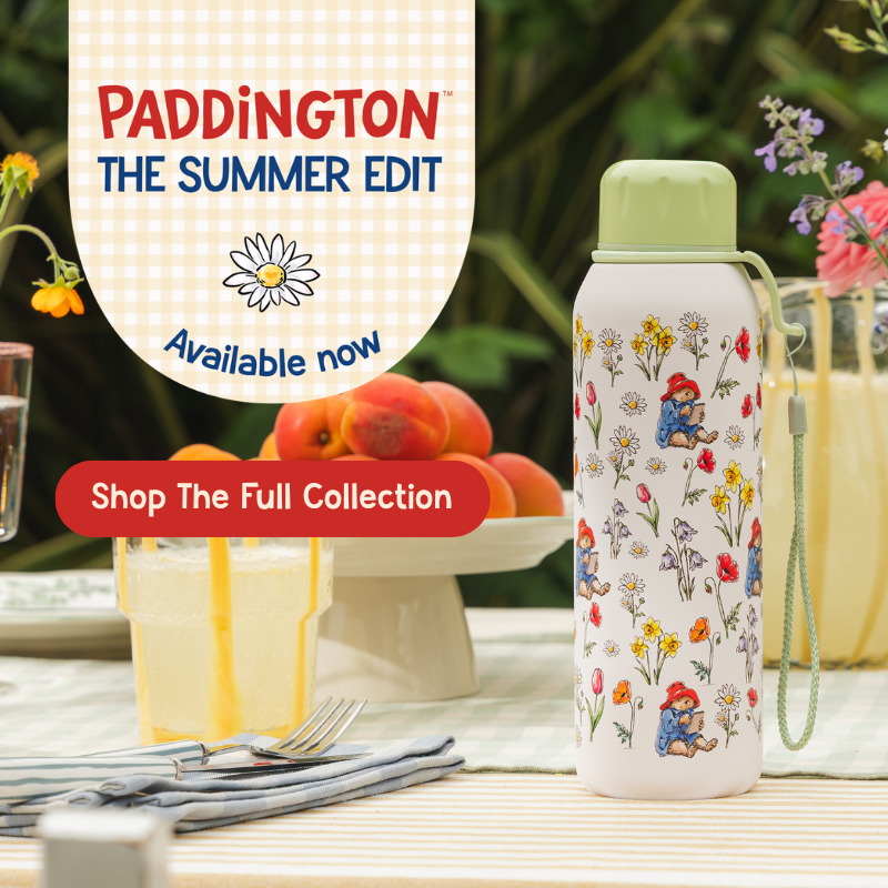 A white insulated water bottle with a light green cap and wrist strap, decorated with a colorful Paddington bear floral print, displayed on a table next to a drink. The image includes promotional text overlays reading 'PADDINGTON THE SUMMER EDIT', 'Available now', and 'Shop The Full Collection'.