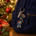 A navy blue backpack with decorative Christmas-themed metal keychain charms, including a 'Paddington' tag, a marmalade jar, a candy cane and a Paddington bear with gifts character charm, shown beside a wrapped gift and a lit Christmas tree.