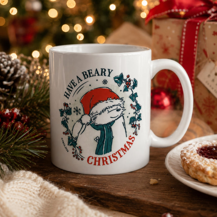A white ceramic mug with a festive Grinch-themed Christmas print, styled on a wooden table with a blurred Christmas tree, gift boxes, and a cookie in the background.