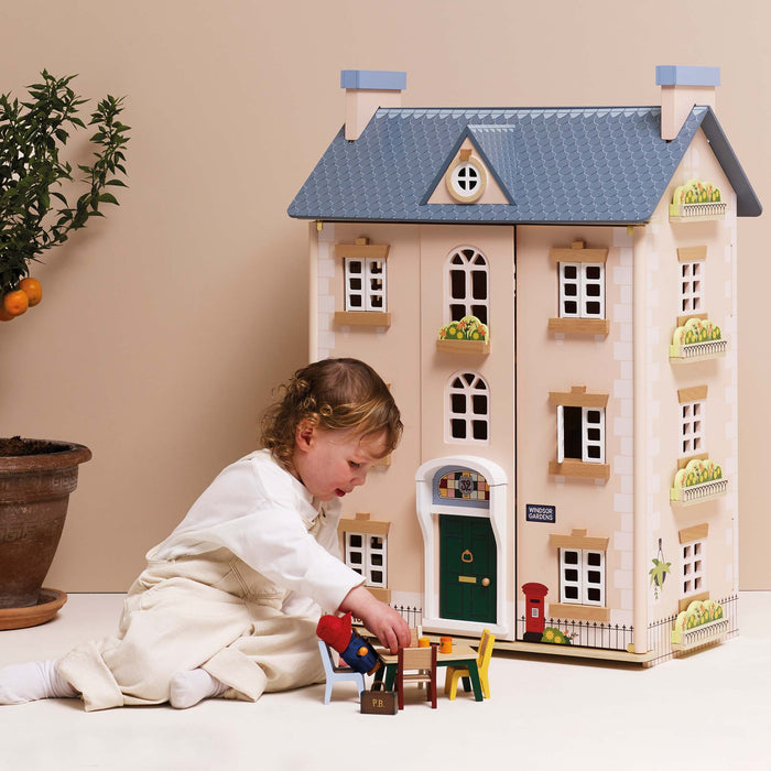 A young child in a white outfit plays with small toy furniture in front of a large wooden dollhouse styled like a house, set against a warm beige background.