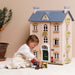 A young child in a white outfit plays with small toy furniture in front of a large wooden dollhouse styled like a house, set against a warm beige background.