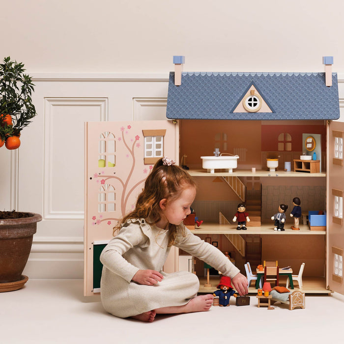 A young girl in a cream-colored dress sits on the floor playing with a large wooden dollhouse. The dollhouse features a blue textured roof, pastel-colored doors with a tree design, and multiple interior rooms with miniature furniture and figures.