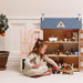 A young girl in a cream-colored dress sits on the floor playing with a large wooden dollhouse. The dollhouse features a blue textured roof, pastel-colored doors with a tree design, and multiple interior rooms with miniature furniture and figures.