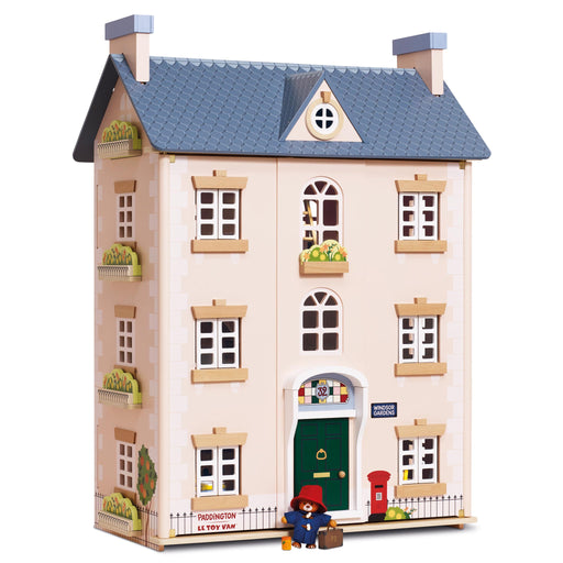A wooden dollhouse-style house with a blue-gray tiled roof, cream-colored walls, multiple windows, a green front door, and small decorative figures and plants, shown on a plain white background.
