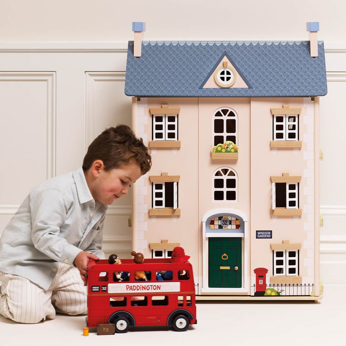A young boy in a light gray shirt and striped pants kneels on the floor next to a red wooden double-decker bus, playing with it in front of a large beige toy house with a blue roof.