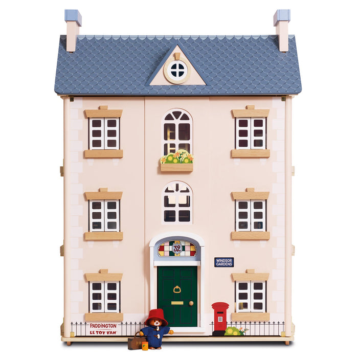 A wooden dollhouse of a three-story house with a blue tiled roof, cream-colored walls, and a green front door. The dollhouse is decorated with small accessories including a Paddington Bear figure, a red post box, and a fruit window box, set against a plain white background.