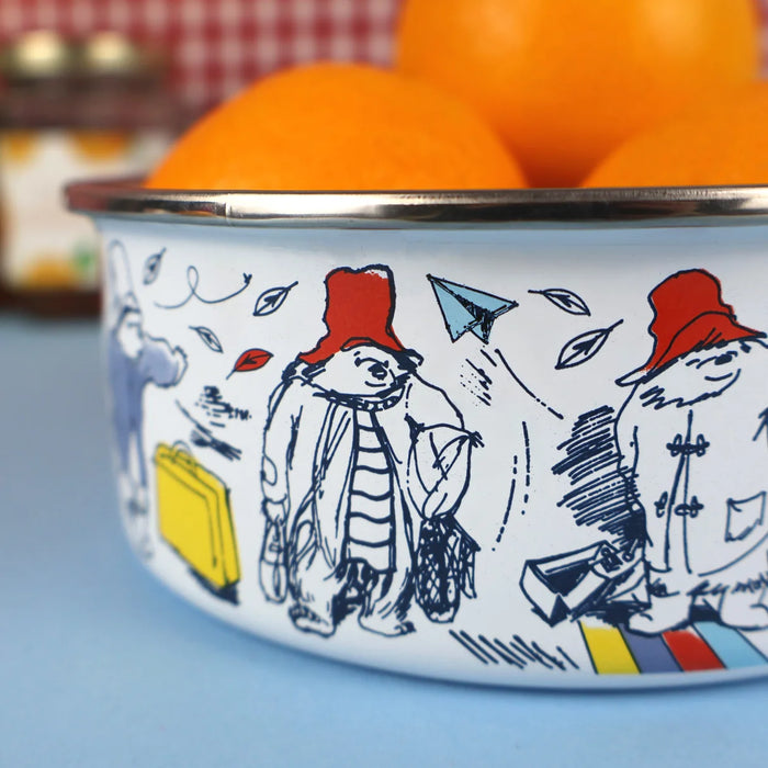 A white enamel bowl with a silver rim, decorated with black line art illustrations of Paddington bear in red hats and a blue paper airplane, filled with oranges on a blue surface.