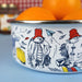 A white enamel bowl with a silver rim, decorated with black line art illustrations of Paddington bear in red hats and a blue paper airplane, filled with oranges on a blue surface.