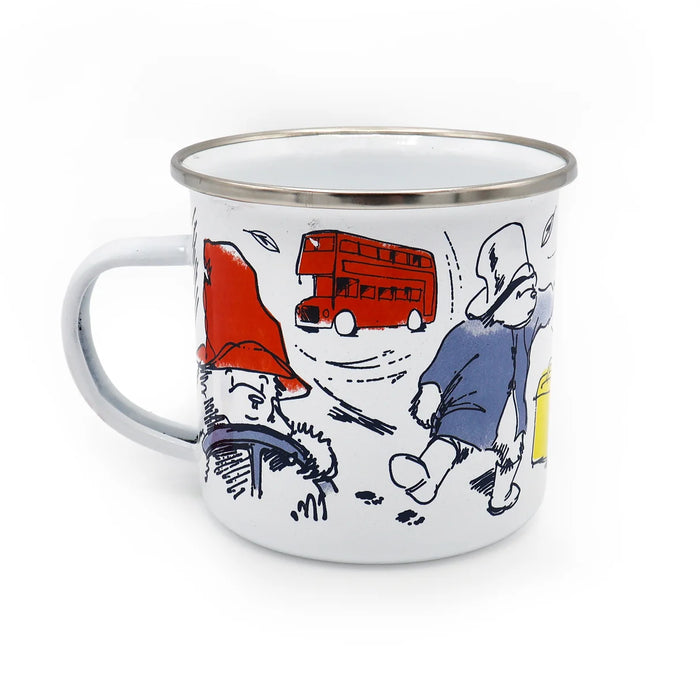 A white enamel camp mug with a silver metal rim, featuring a whimsical cartoon paddington bear illustration in a red hat, a red double-decker bus, and a bear figure in blue.