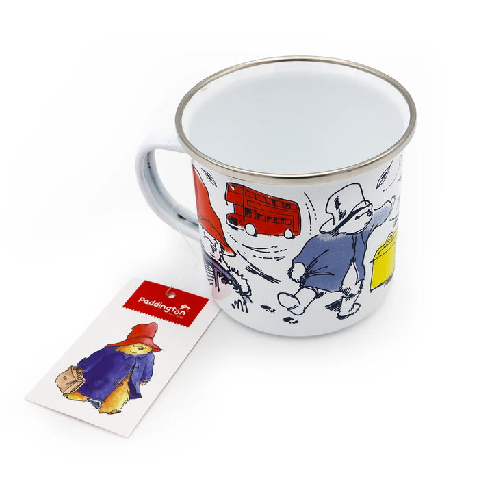 A white enamel camping mug with a silver rim, featuring a colorful illustration of Paddington Bear walking alongside a red double-decker bus. A small printed card with Paddington branding is placed in front of the mug on a clean white background.