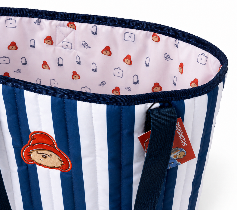 A corner shot of a navy blue and white striped quilted Paddington beach bag with Paddington bear face logo embroidery, navy blue handles and Paddingonton-esque printed lining.