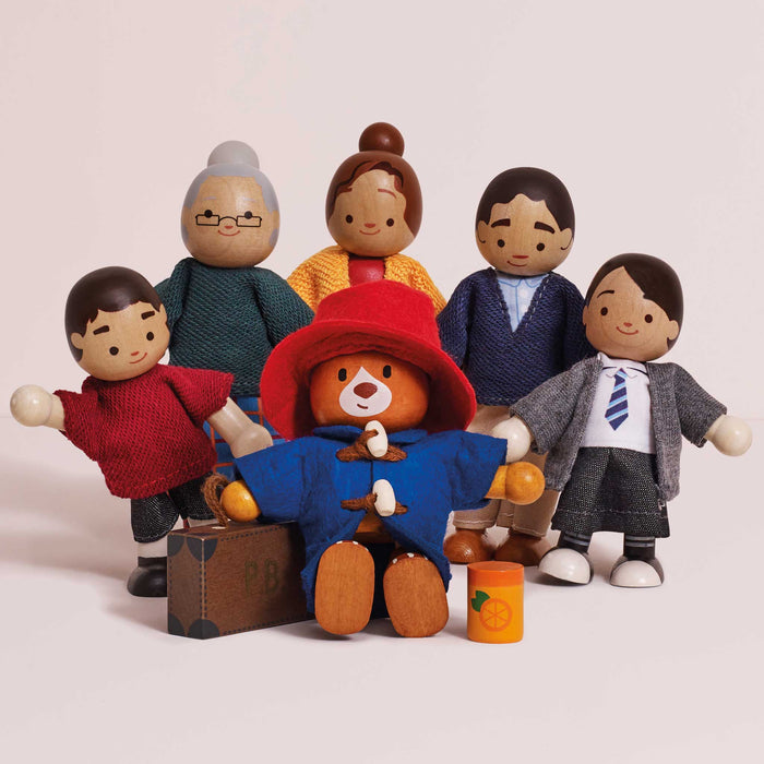 A set of wooden toy figures and accessories arranged together on a plain light background. The set includes several child and adult figures in different colored outfits, Paddington bear figure wearing a red hat, a brown suitcase, and an orange jar.