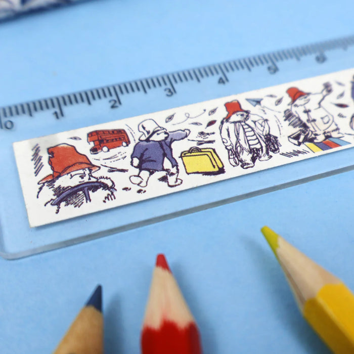 A close-up of a white ruler featuring Paddington bear characters and objects, laid diagonally on a light blue background with coloured pencil tips nearby.