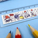 A close-up of a white ruler featuring Paddington bear characters and objects, laid diagonally on a light blue background with coloured pencil tips nearby.