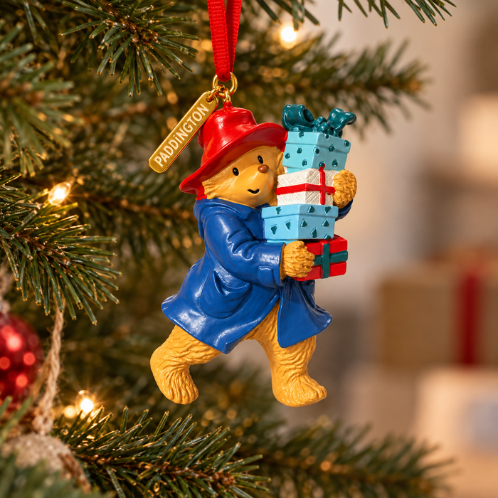 A Paddington Christmas ornament hanging from a Christmas tree, wearing a red hat and blue coat while carrying wrapped presents. The ornament has a red hanging ribbon and a gold tag that reads 'PADDINGTON'.