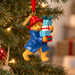 A Paddington Christmas ornament hanging from a Christmas tree, wearing a red hat and blue coat while carrying wrapped presents. The ornament has a red hanging ribbon and a gold tag that reads 'PADDINGTON'.