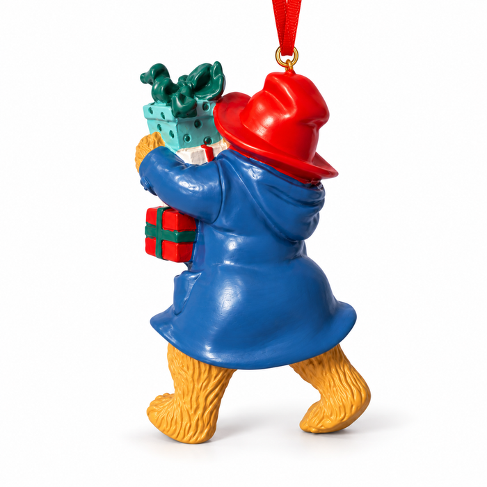 A Christmas ornament shaped like Paddington Bear carrying a gift box, featuring a blue coat, red hat, yellow fur, and a red hanging ribbon on a white background.