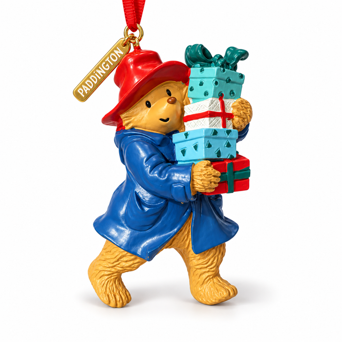 A Paddington bear Christmas ornament featuring the bear in his signature blue coat and red hat, carrying a stack of wrapped presents, with a red hanging ribbon and a gold tag reading PADDINGTON.