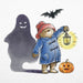 Illustration of paddington bear character in a blue coat with a lantern, ghost, bat, and pumpkin on a white background
