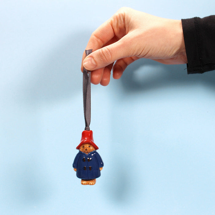 Hand holding a christmas decoration with a Paddington Bear figure against a light blue background