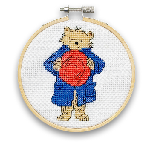 A mini cross stitch kit featuring Paddington Bear with a red hat and a blue coat on a white fabric hoop.