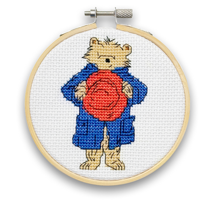 A mini cross stitch kit featuring Paddington Bear with a red hat and a blue coat on a white fabric hoop.