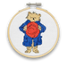 A mini cross stitch kit featuring Paddington Bear with a red hat and a blue coat on a white fabric hoop.