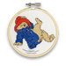 A mini cross stitch kit featuring an image of Paddington Bear wearing a blue outfit, falling over, with a white background.