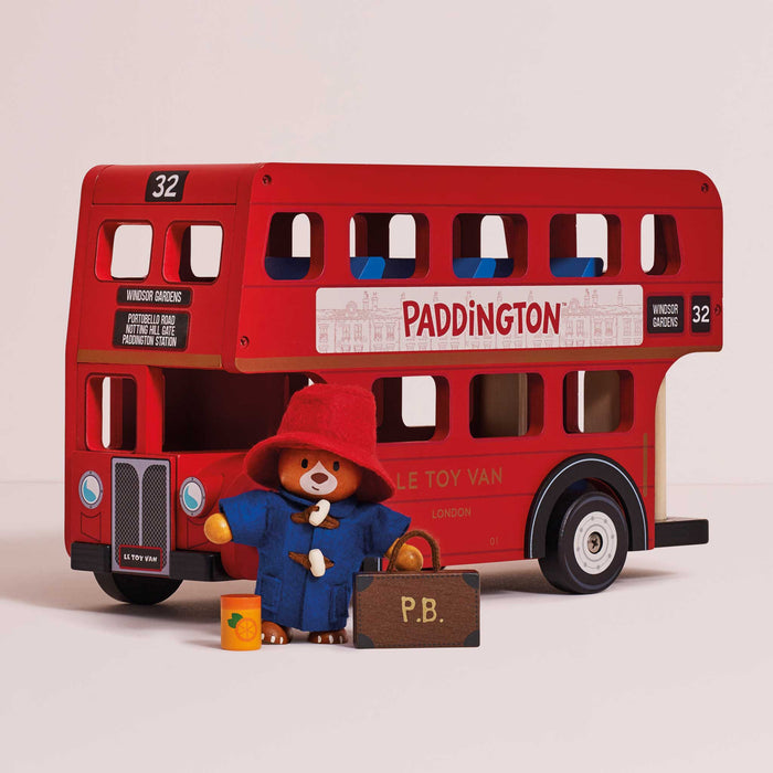A red double-decker toy bus with Paddington Bear figurine and suitcase in front, photographed on a clean light beige studio background.