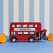 A red double-decker toy bus with Paddington Bear seated at the rear, set against a blue and white striped backdrop with orange slices on both sides.