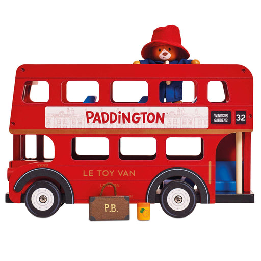 A wooden toy double-decker bus styled like a red London bus, with Paddington Bear visible inside behind a red hat and blue coat. The side of the bus reads “PADDINGTON” and “WINDSOR GARDENS,” with “LE TOY VAN” on the front, and a small brown suitcase and yellow bottle placed in front on a white background.