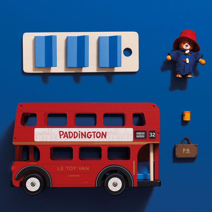 A wooden toy double-decker bus set on a blue background, including a red bus labeled Paddington, a cream bus panel with blue window pieces, a Paddington bear figure in a red hat and blue coat, a small yellow cup, and a brown briefcase.