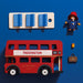 A wooden toy double-decker bus set on a blue background, including a red bus labeled Paddington, a cream bus panel with blue window pieces, a Paddington bear figure in a red hat and blue coat, a small yellow cup, and a brown briefcase.