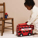 A young child in a cream outfit pushes a red double-decker toy bus labeled PADDINGTON. A wooden chair on the left holds a small Paddington figurine against a plain beige studio background.